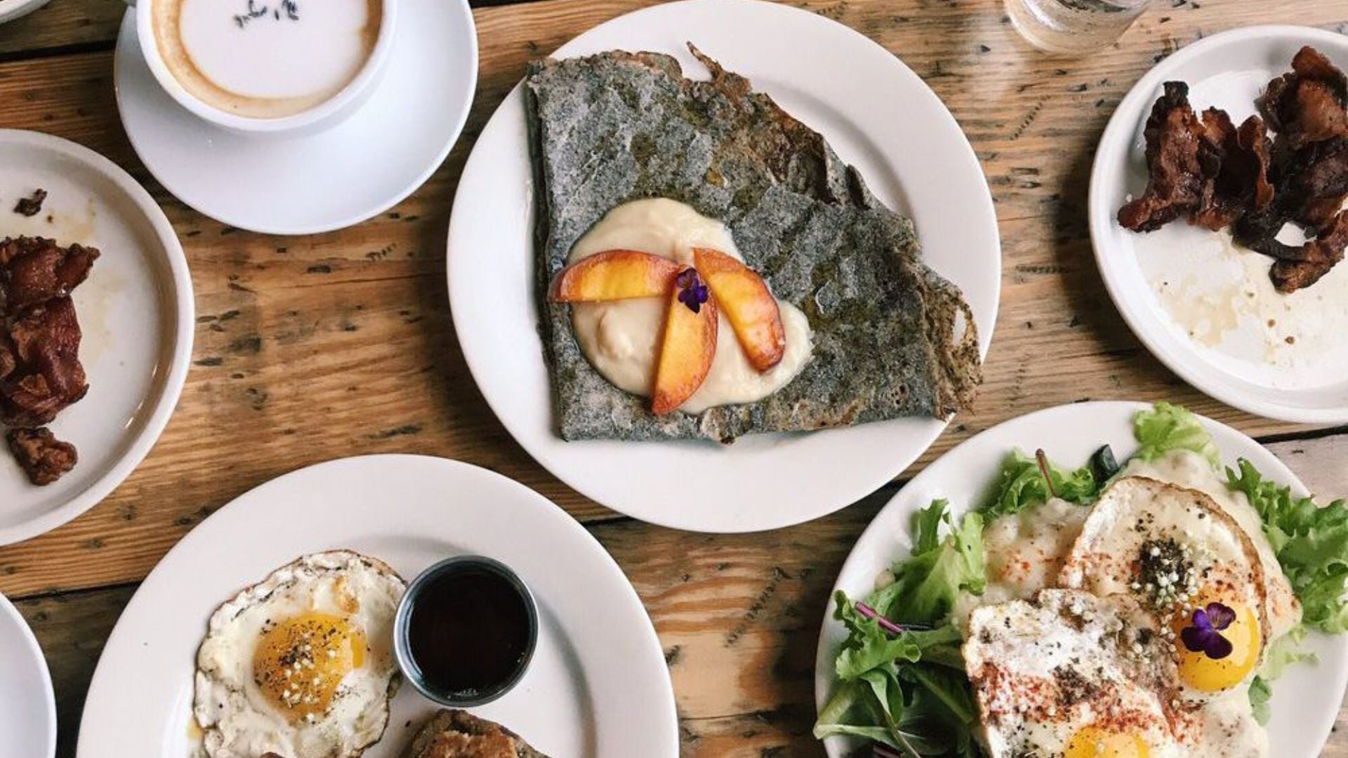 A rustic wooden table set with plates of fried eggs, bacon, a buckwheat crepe topped with fruit and cream, salad greens, toast with syrup, and a cup of coffee with foam art—just like you'd find at beloved under-the-radar restaurants.