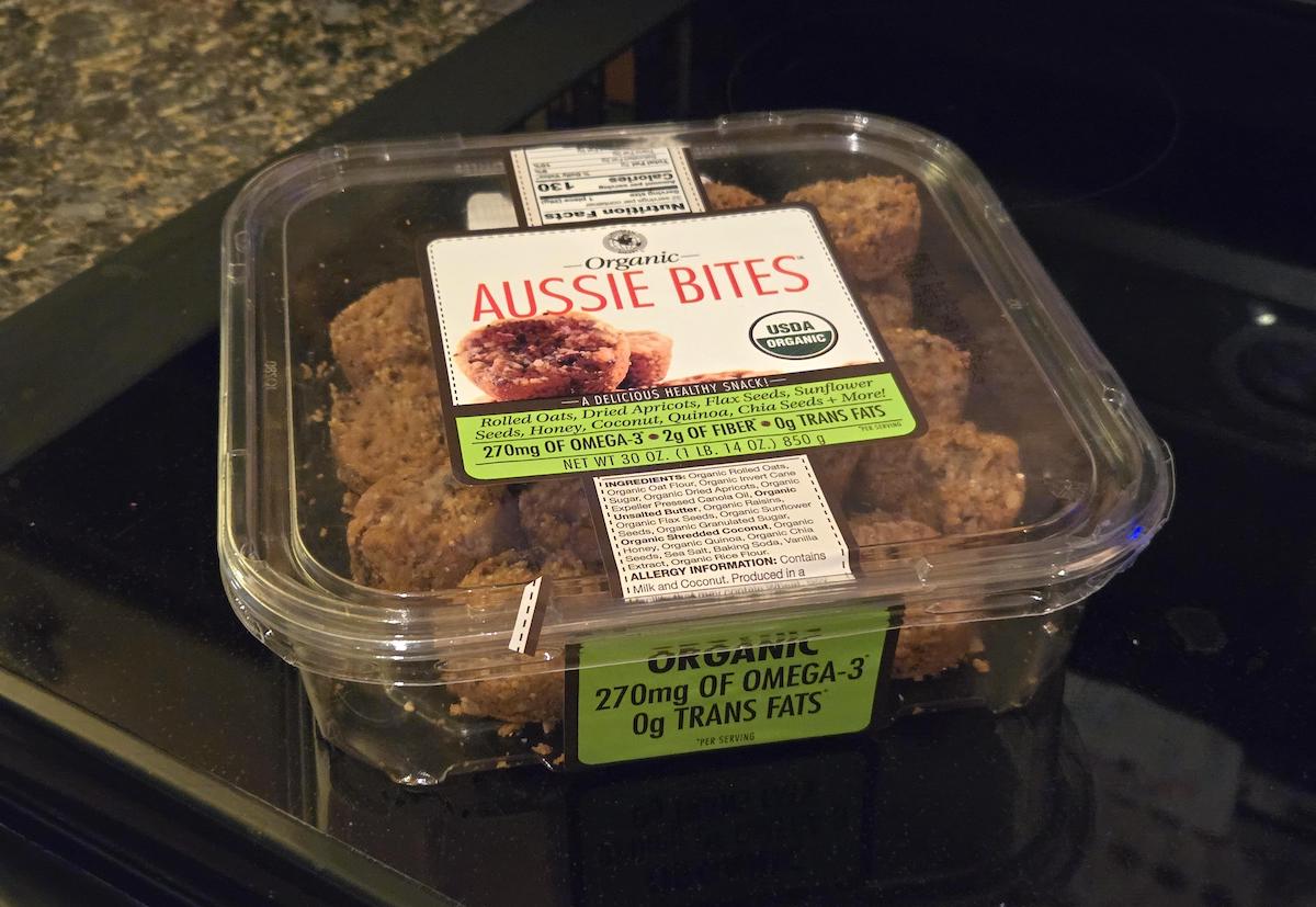 A clear plastic container of Organic Aussie Bites sits on a kitchen counter, showing bite-sized snacks inside. The label highlights 270mg Omega-3 and 0g trans fats, with USDA Organic certification.