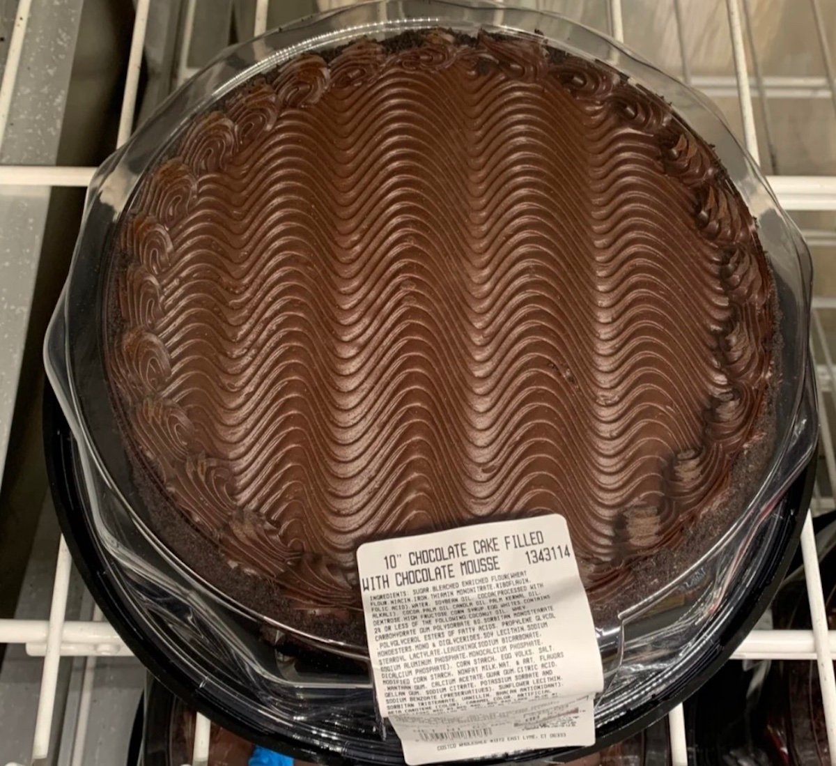 A round chocolate cake with chocolate mousse filling, topped with decorative wavy swirls of chocolate frosting, sits in a clear plastic container with a label on top.
