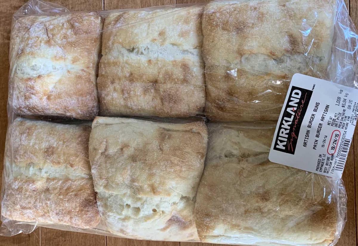 A package of six Kirkland brand square ciabatta sandwich rolls in clear plastic wrapping, showing the golden-brown crusty tops and a white product label on the right side.