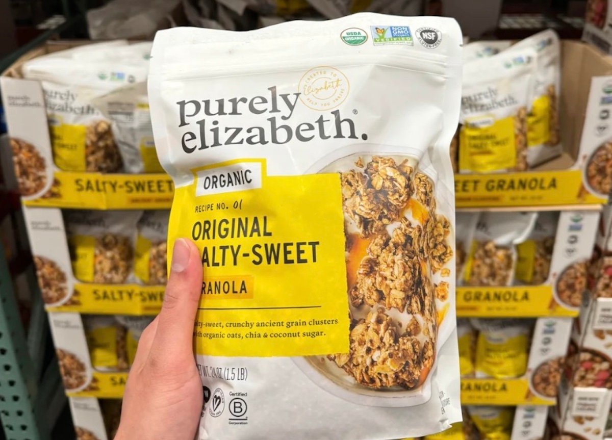 A hand holds a bag of Purely Elizabeth Organic Original Salty-Sweet Granola in front of a store shelf stocked with similar bags. The packaging is mostly white with a large yellow label.