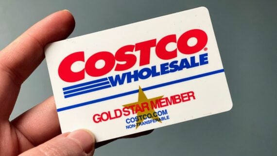 A hand holds a Costco Wholesale membership card labeled "Gold Star Member" with red and blue text, a yellow star, and the Costco website address on a white background.