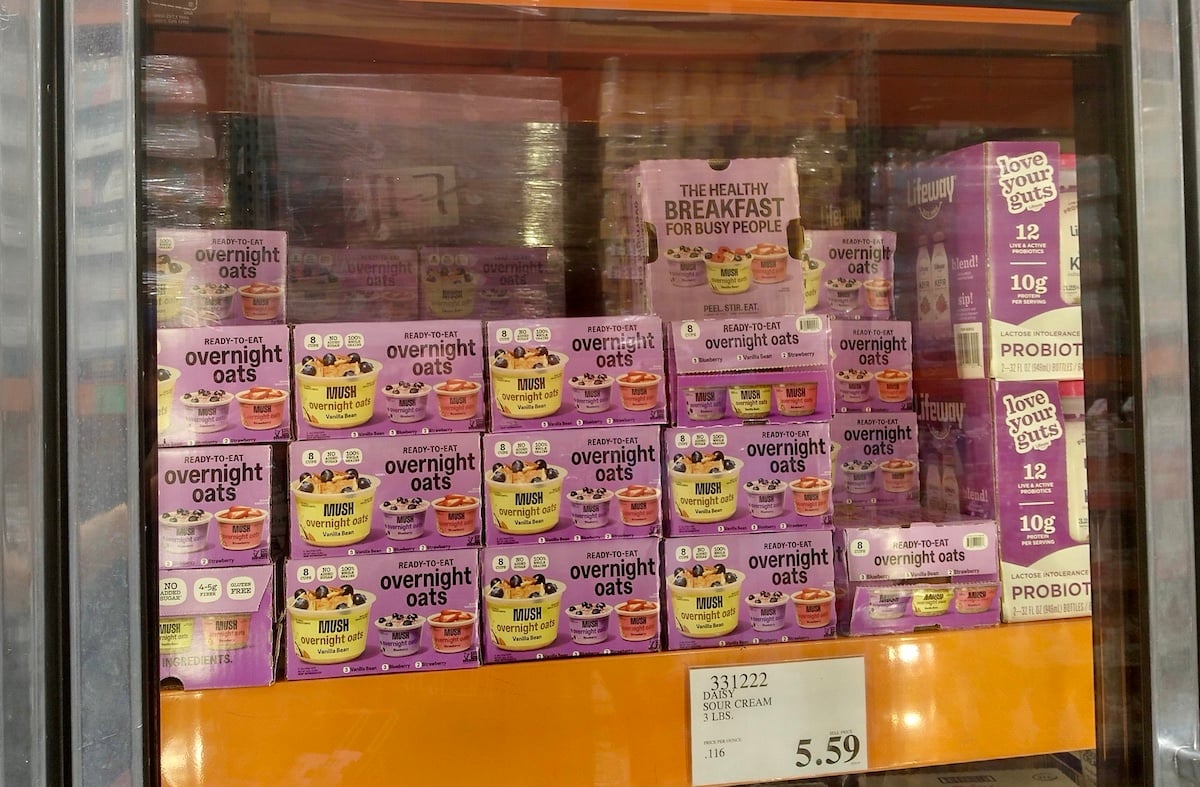 Multiple boxes of "overníght oats" in purple packaging are stacked on a refrigerated shelf in a store, with a price tag below showing $5.59. A sign on the shelf reads, “THE HEALTHY BREAKFAST FOR BUSY PEOPLE.”