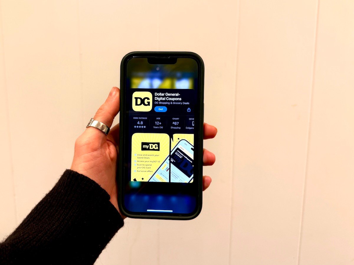 A hand holds a smartphone displaying the Dollar General app page in the App Store, showing the app’s logo, ratings, and install button on the screen. The background is plain and light-colored.