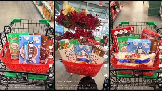 A red gift basket filled with snacks like Swiss Miss, Funfetti, and cookies sits in a shopping cart at a Dollar Tree store. The basket is surrounded by store aisles and red artificial flowers.