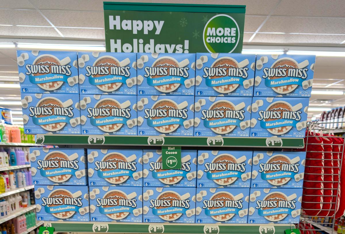 A store shelf displays multiple boxes of Swiss Miss Marshmallow hot cocoa mix under a green sign that reads "Happy Holidays! More Choices." The boxes are neatly stacked in rows.