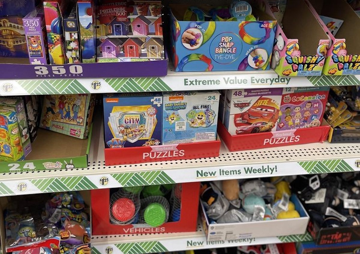 Store shelves display a variety of colorful puzzles, games, and toys, including puzzles with cartoon characters and small boxed toys, all marked with signs reading “Extreme Value Everyday” and “New Items Weekly.”