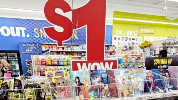 A retail store display with a large red "$1 WOW" sign above bins of coloring books and toys. Shelves in the background hold balloons, snacks, and party supplies. Shoppers browse the brightly lit aisle.