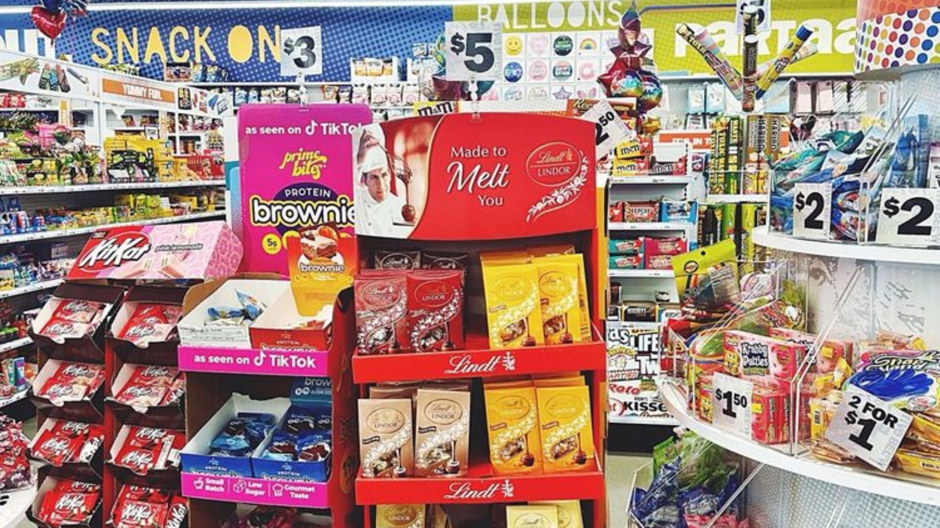 A colorful store aisle displays shelves of snacks, chocolates, and candy. Prominent signs show prices like $2, $3, and $5. Lindt chocolates and protein brownies are featured on central red and purple stands.