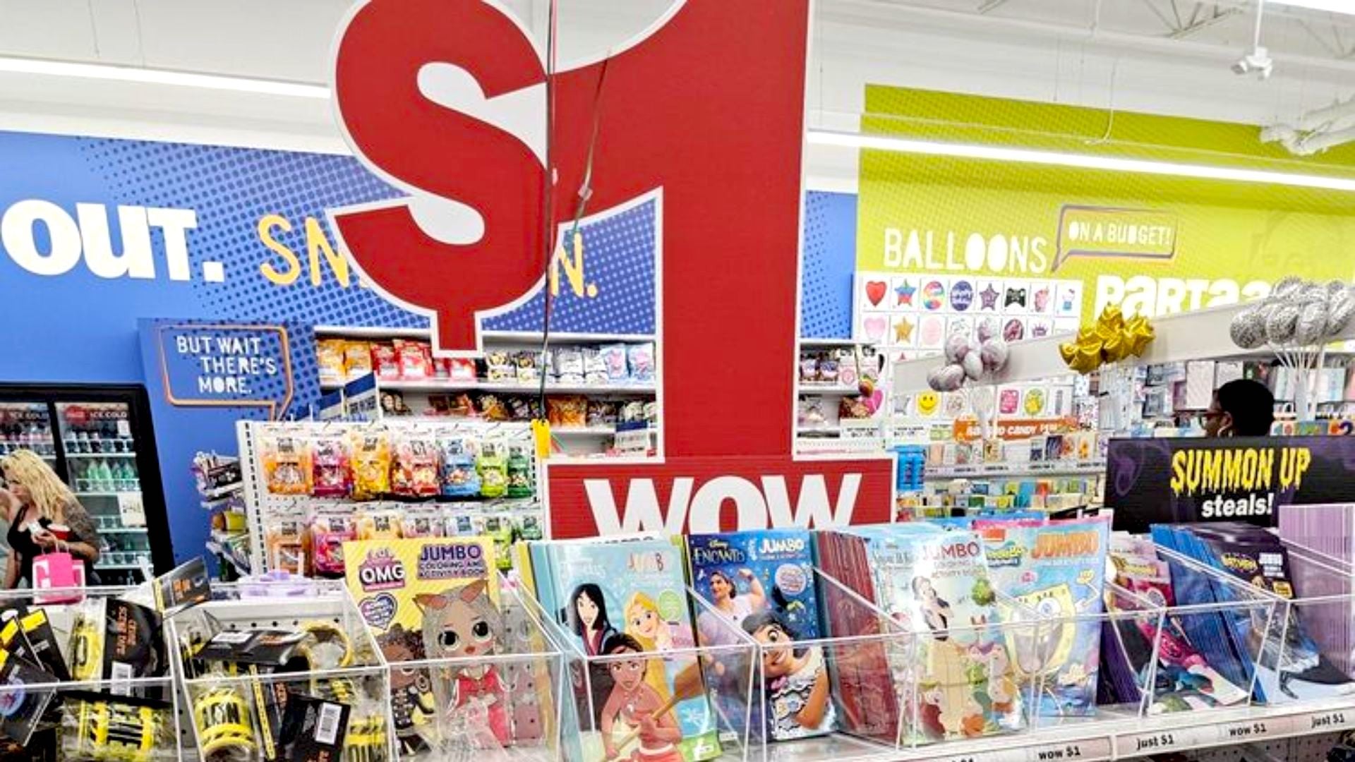 A retail store display with a large red "$1 WOW" sign above bins of coloring books and toys. Shelves in the background hold balloons, snacks, and party supplies. Shoppers browse the brightly lit aisle.