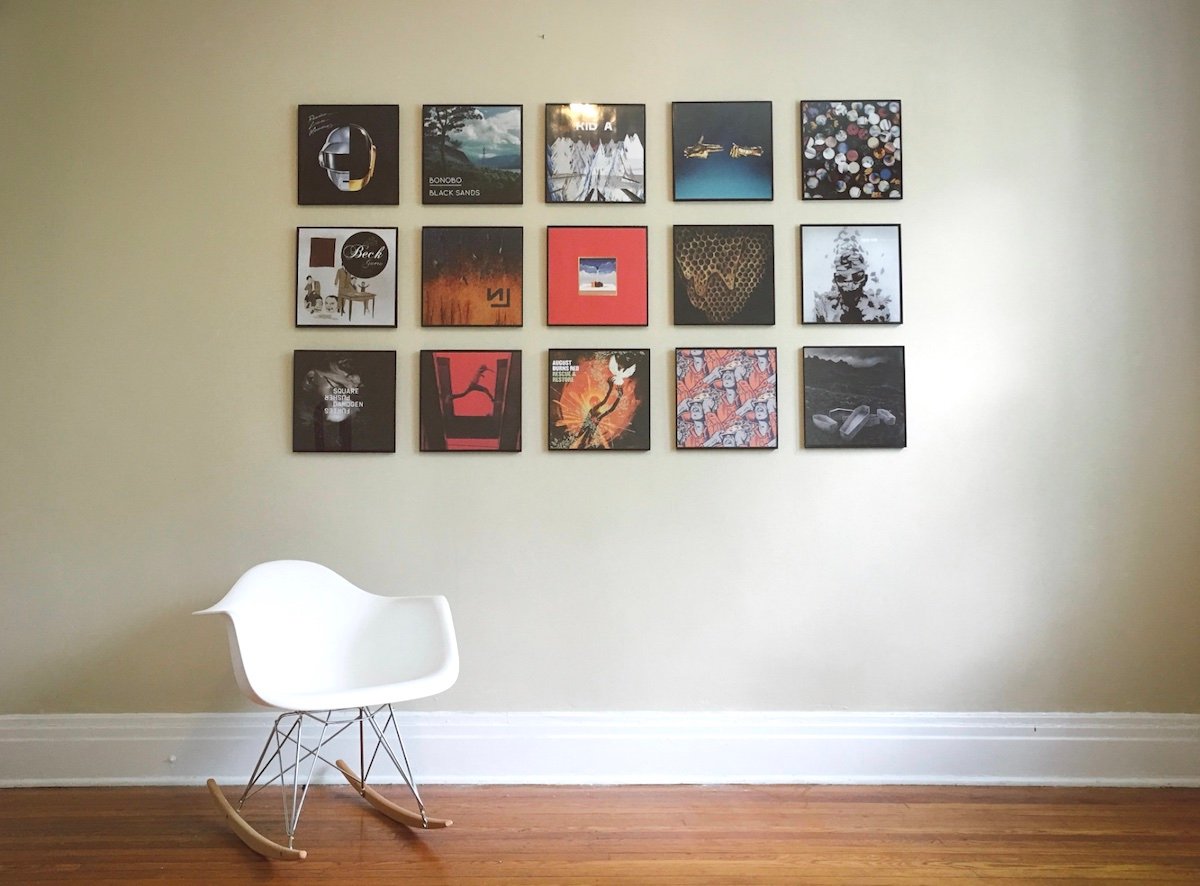 A white rocking chair sits on a wooden floor against a beige wall, above which 15 square album covers are neatly arranged in a 3-by-5 grid.