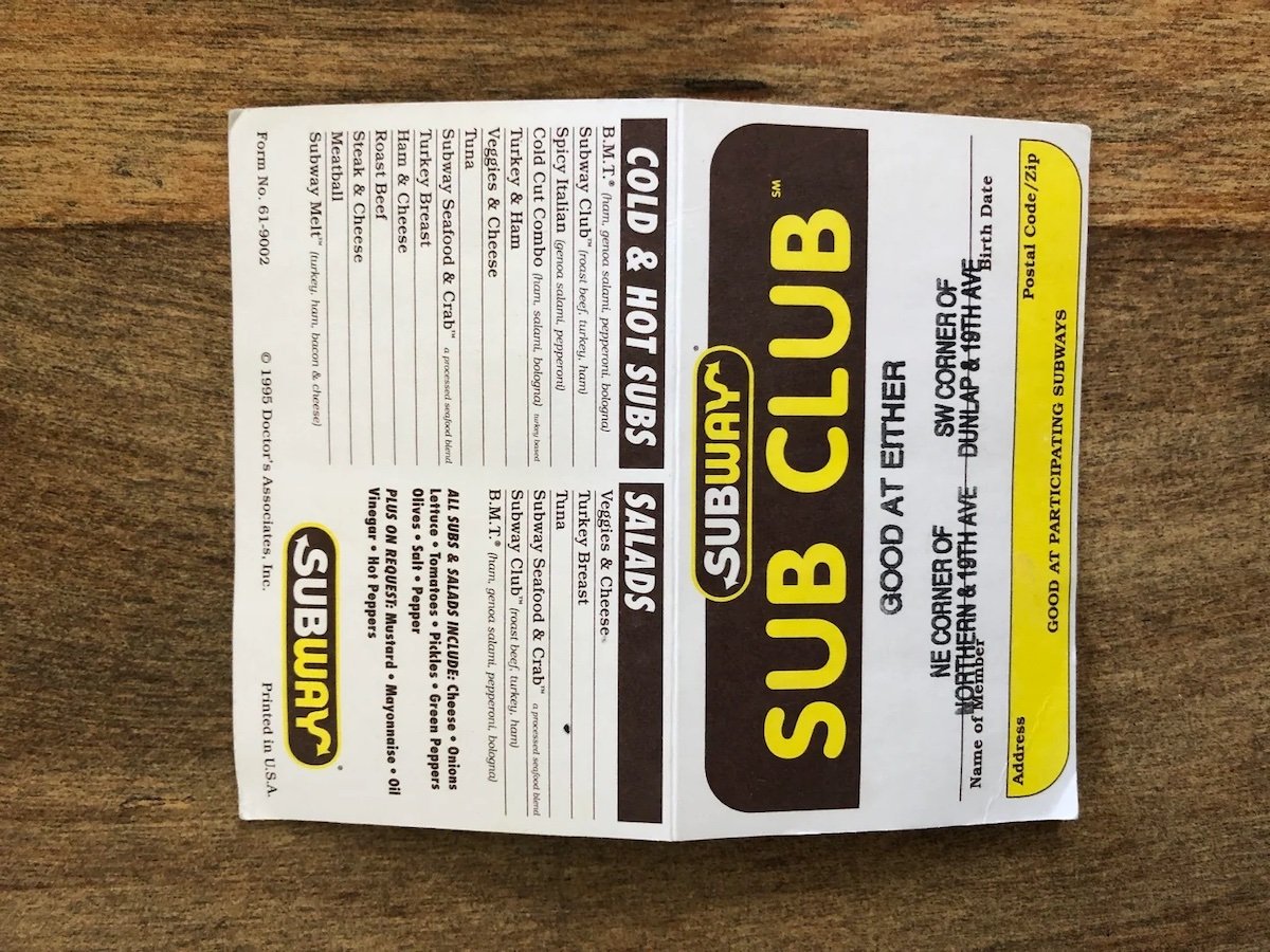 A vintage Subway Sub Club stamp card lies open on a wooden surface, showing spaces for stamps and text for redeeming a free sub at specific Subway locations.