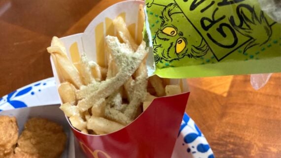 French fries in a red container are being sprinkled with green powder from a packet labeled with a drawing of the Grinch and the words “Grinch Green.” The fries are on a plate next to chicken nuggets.