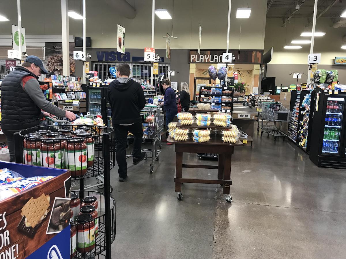 Shoppers stand in line with carts at a grocery store checkout. Shelves display snacks, drinks, and bakery items. Signs and displays are visible, and the store has bright lighting with a visible "PLAYLAND" sign in the background.
