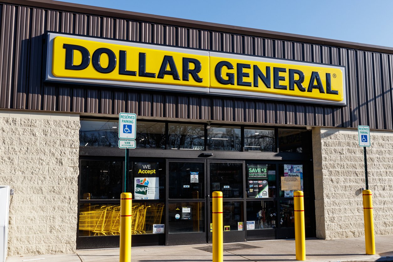 Front entrance of a Dollar General store with a large yellow sign, shopping carts visible inside, and two handicapped parking signs by the doors on a sunny day.