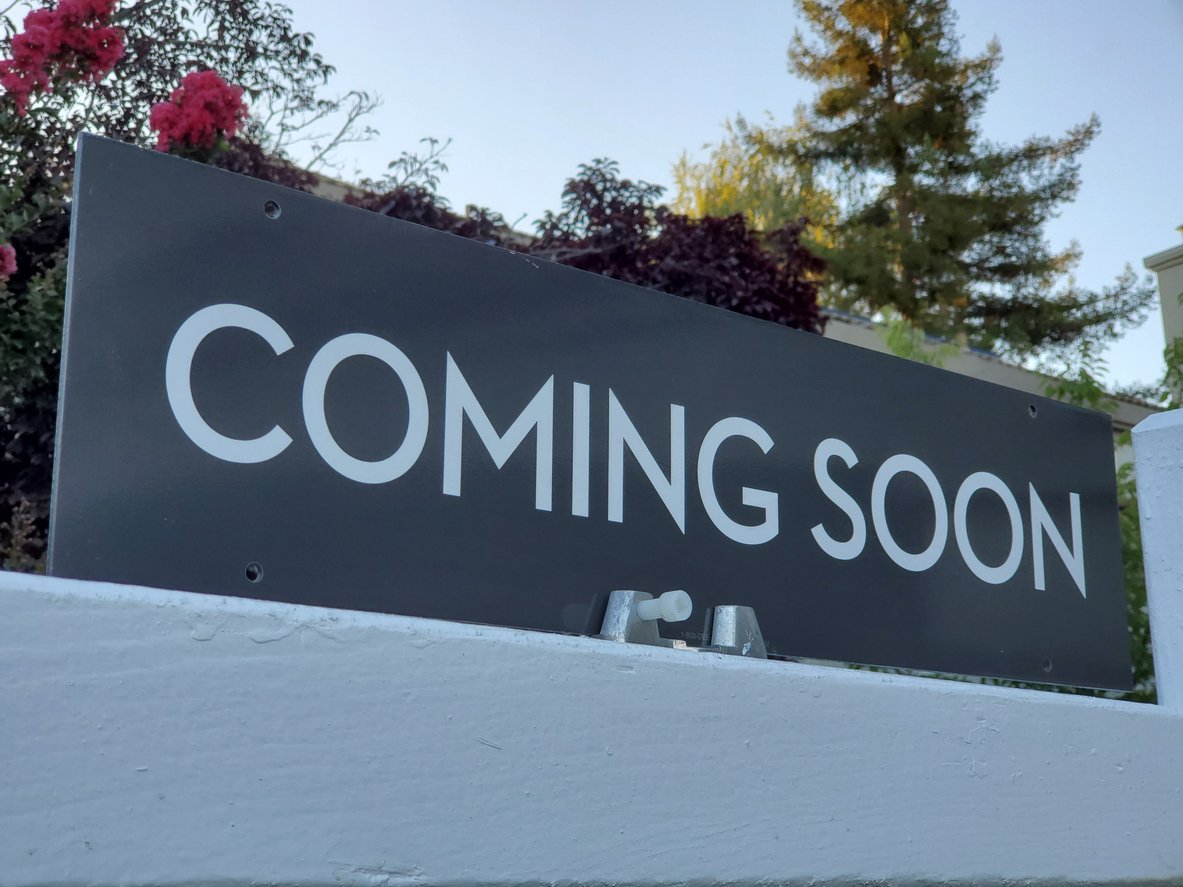 A black sign with large white text that reads "COMING SOON," mounted on a white fence with trees and flowers in the background.