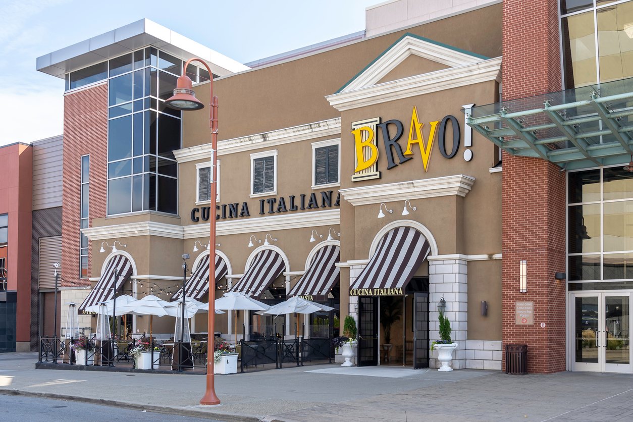 Street view of an Italian restaurant called Bravo! Cucina Italiana, featuring striped awnings, outdoor seating with white umbrellas, and a large yellow and black sign above the entrance.