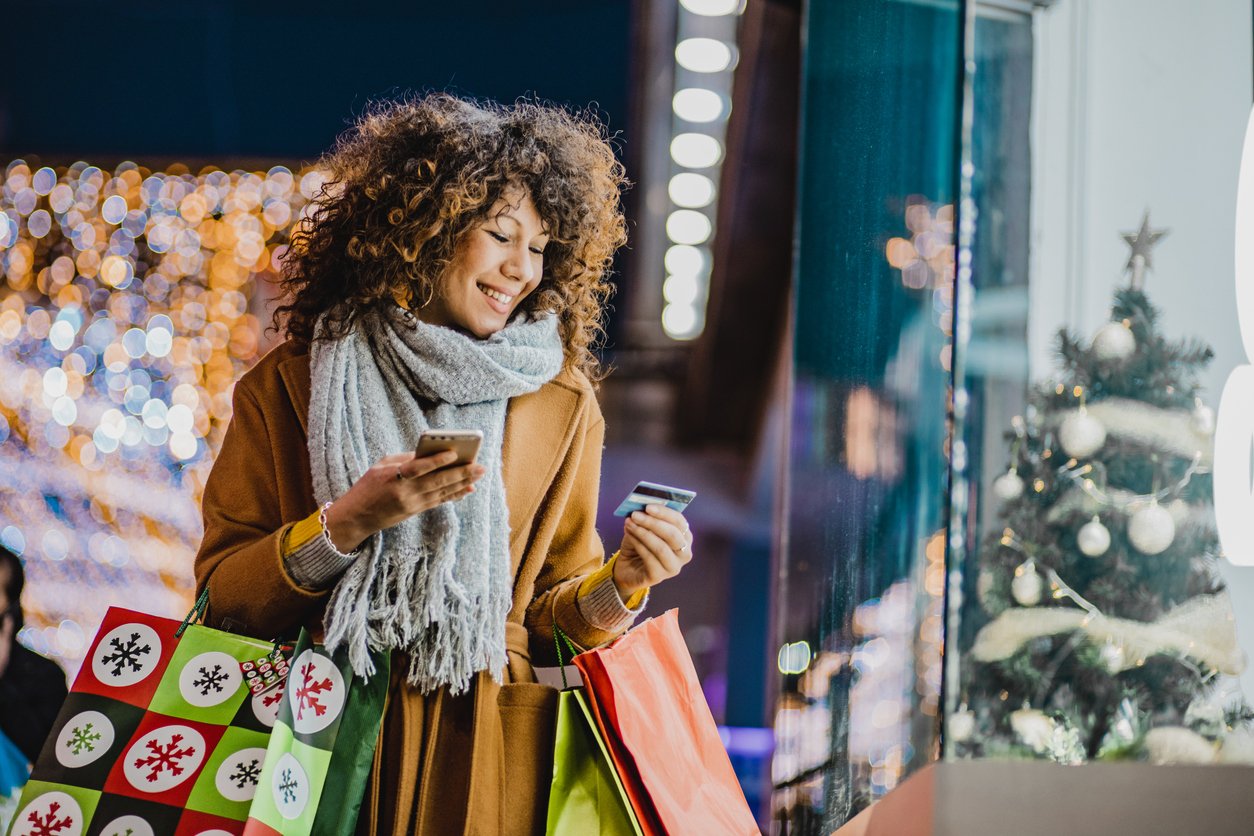 A smiling woman in a coat and scarf holds shopping bags, a smartphone, and a credit card while standing outside a decorated store window with a Christmas tree and festive lights.