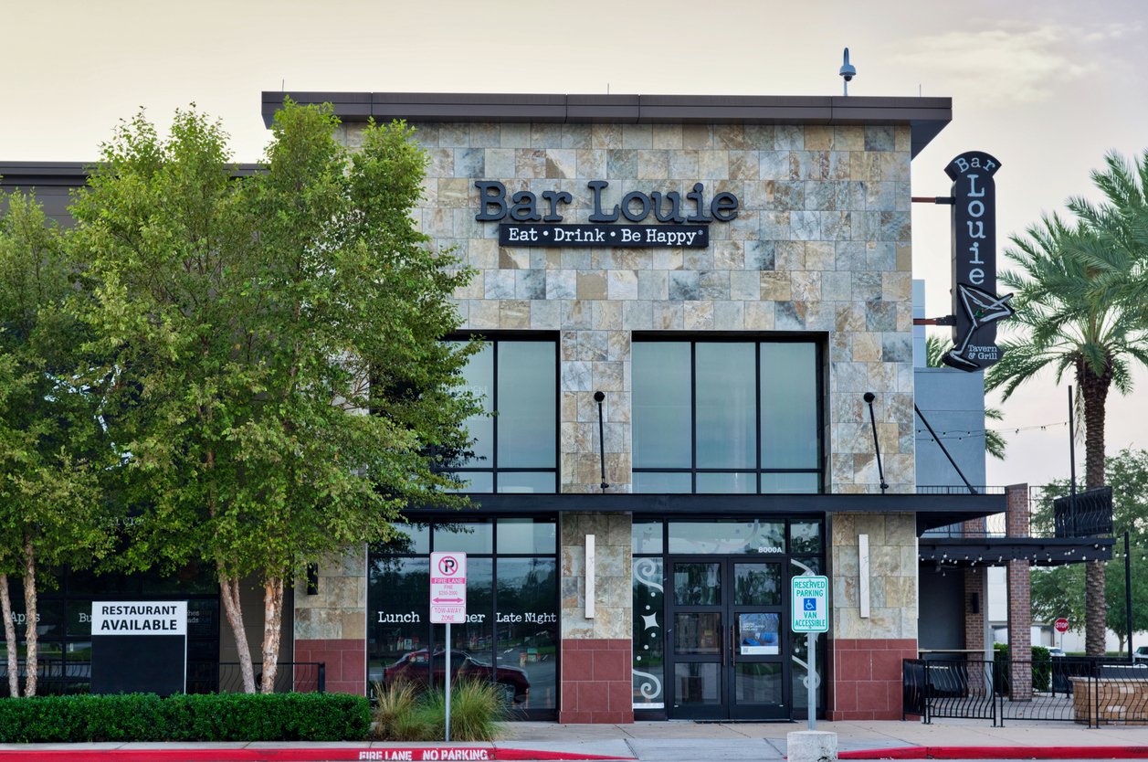 Front view of a Bar Louie restaurant with a stone facade, large windows, trees on both sides, and visible signs for parking and restaurant availability. The text “Eat · Drink · Be Happy” is displayed above the entrance.