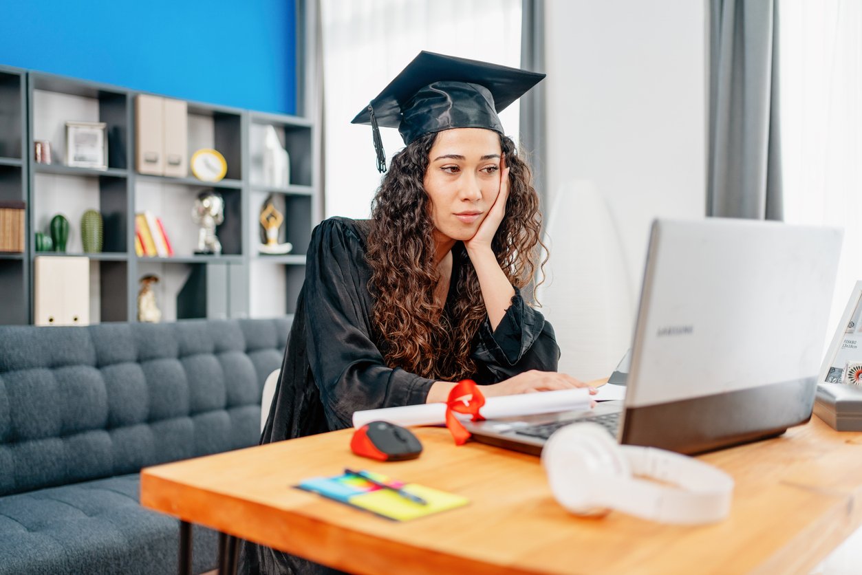 A young woman in a graduation cap and gown sits at a desk, looking at a laptop screen. She appears thoughtful and is resting her face on her hand. The room is modern with shelves, books, and natural light.