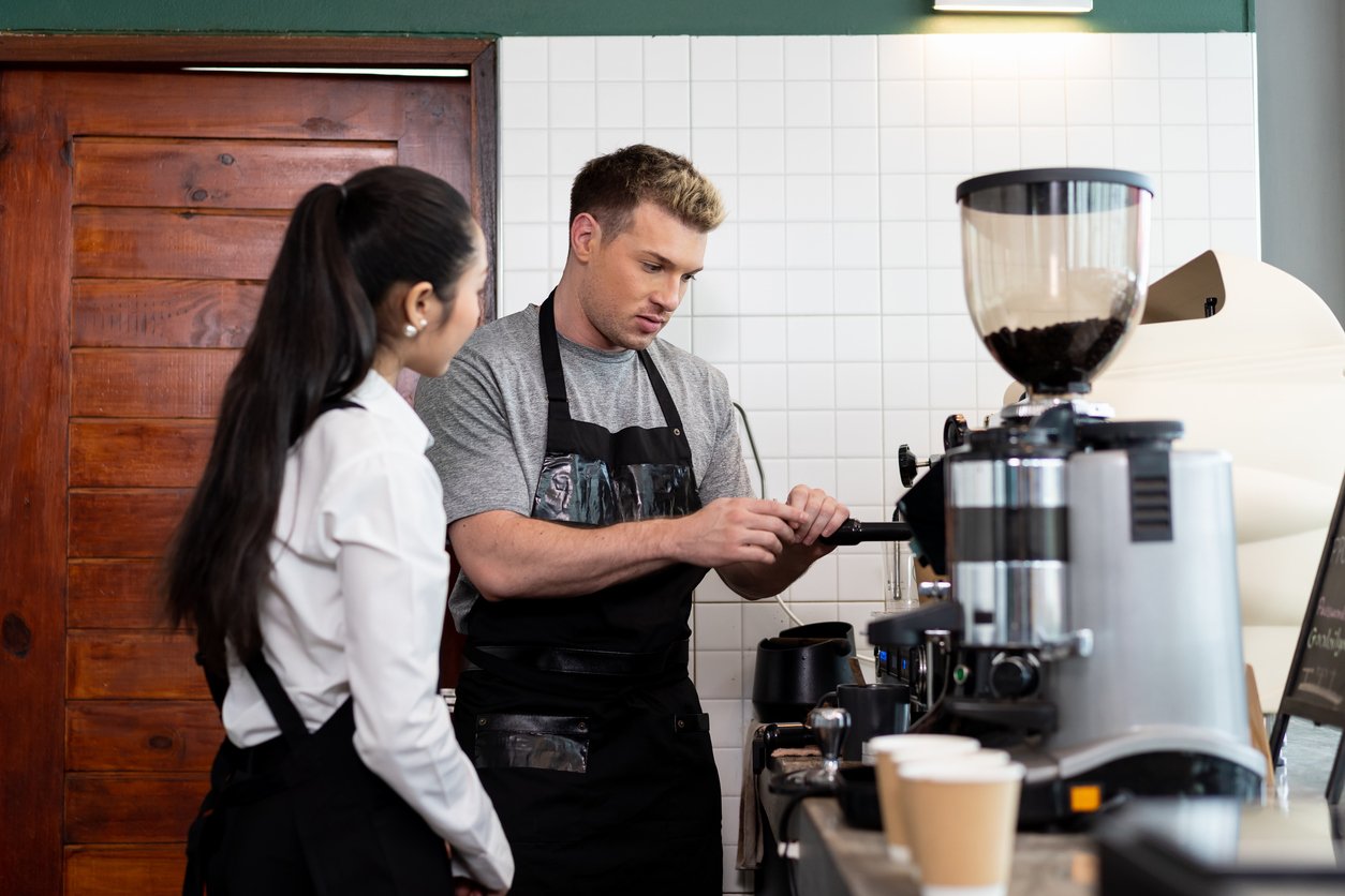 A barista demonstrates how to use an espresso machine to a colleague in a coffee shop. They both wear aprons, and disposable cups are visible on the counter in front of them.