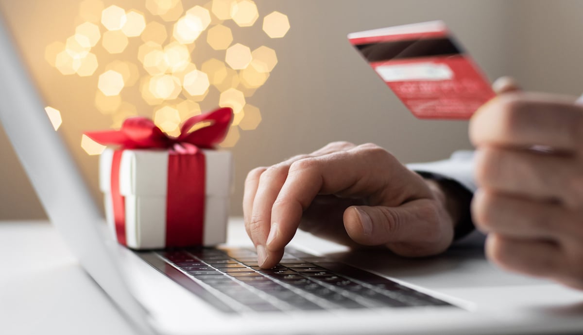 A person uses a laptop while holding a credit card. Next to the laptop is a small, wrapped gift box with a red ribbon. Blurred festive lights appear in the background.