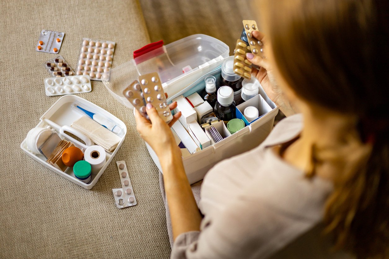 A woman sits on a beige surface, organizing a first aid kit filled with pill blister packs, bottles, bandages, and medical supplies. She holds medication strips in both hands, examining them closely.