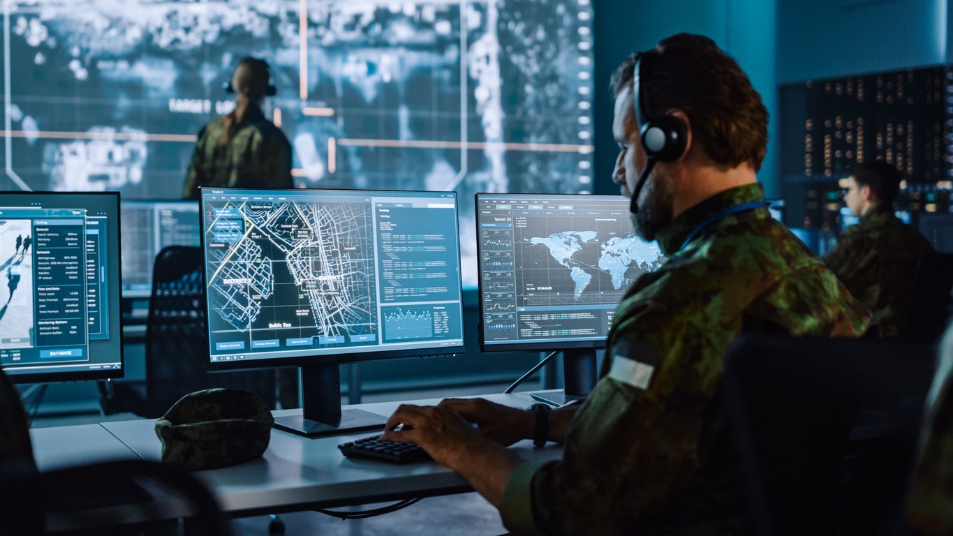 A person in military uniform sits at a desk with multiple computer monitors displaying maps, data, and satellite images in a high-tech control room, with others working in the background.