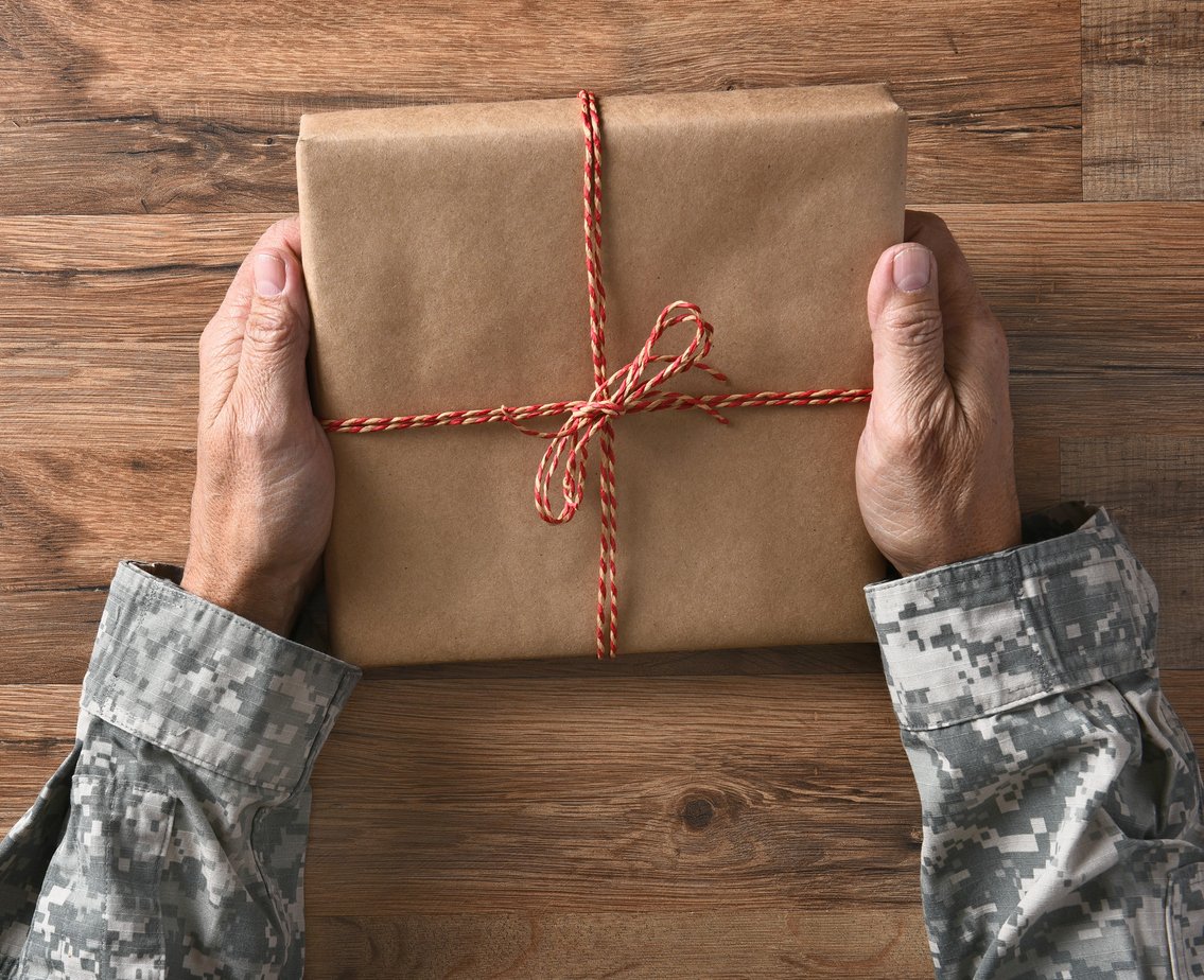 A person in a military uniform holds a gift wrapped in brown paper and tied with a red and white string, placed on a wooden surface.
