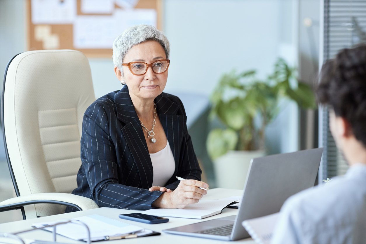A woman with short gray hair and glasses, wearing a striped blazer, sits at a desk with papers, a phone, and a laptop, attentively listening to someone across from her in a modern office.