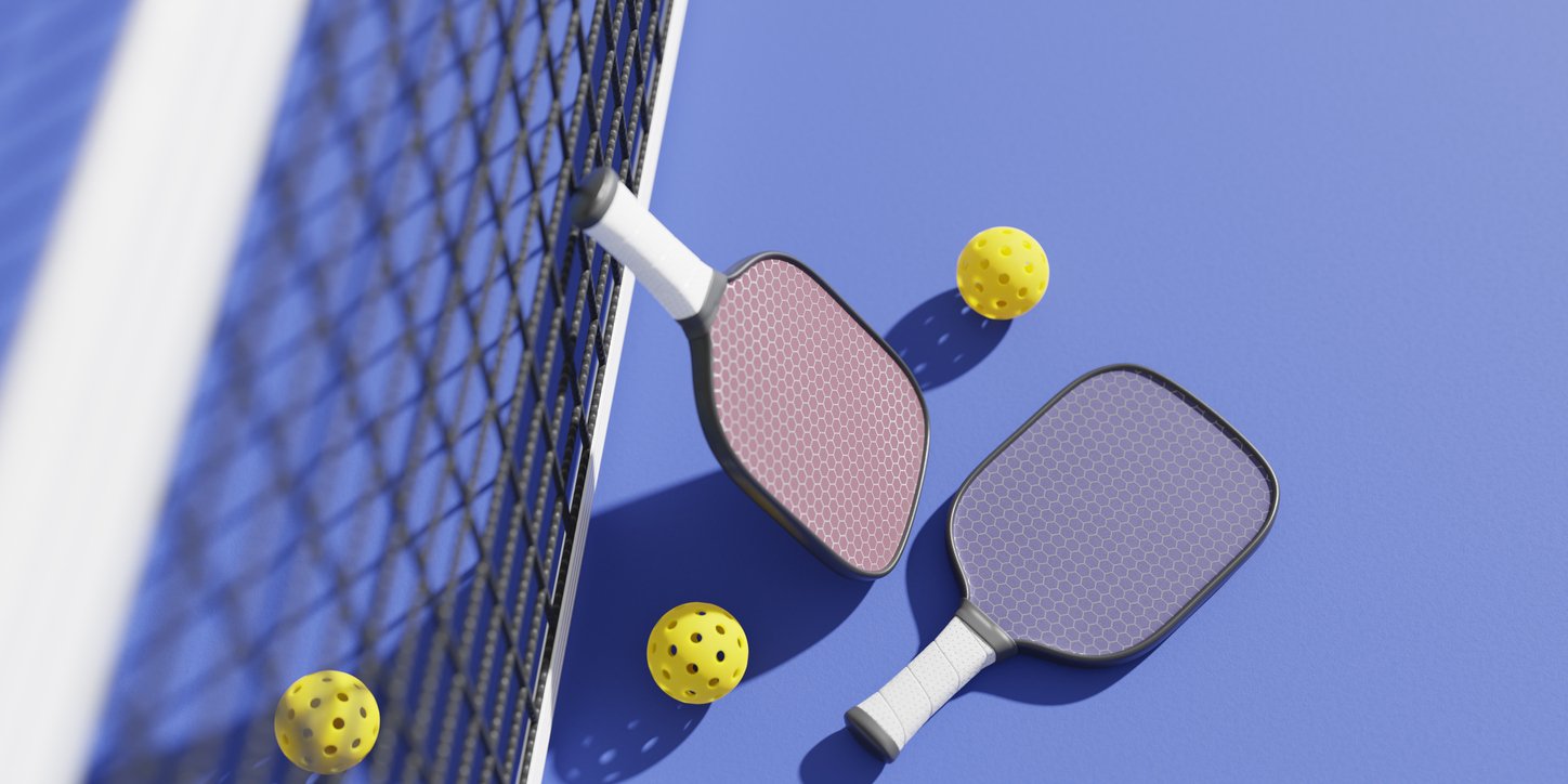 Two pickleball paddles and three yellow perforated balls lie on a blue court next to a black net, shown from an overhead perspective.