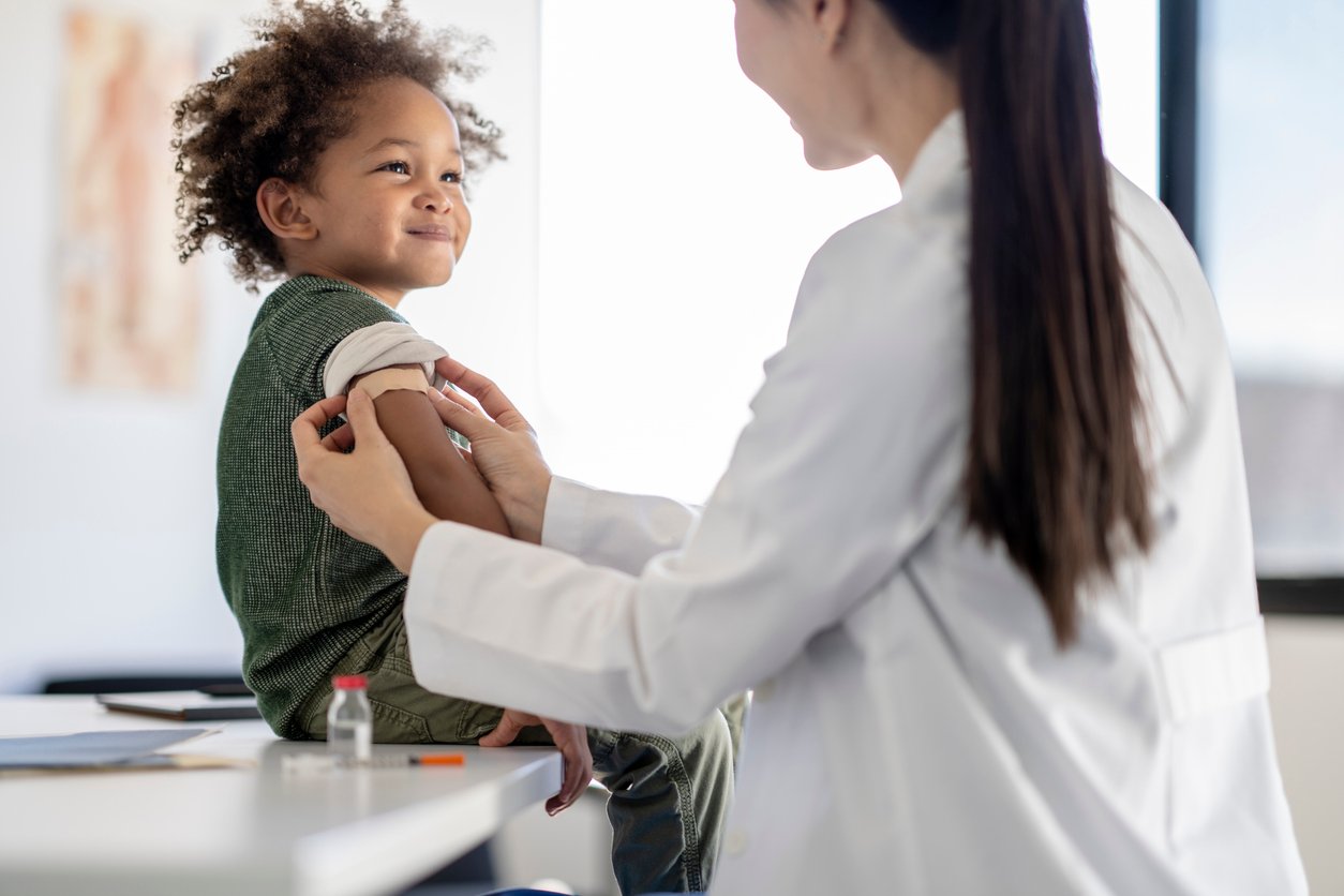 A smiling child sits on an exam table while a healthcare professional in a white coat gently rolls up the child’s sleeve, preparing for a vaccination. A vaccine vial and syringe are visible on the table nearby.