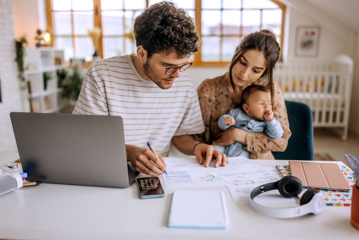 A young couple sits at a desk with a laptop, papers, and calculator, reviewing documents. The woman holds a baby, while the man writes on a form. Headphones and a notebook are also on the desk in a bright, modern room.