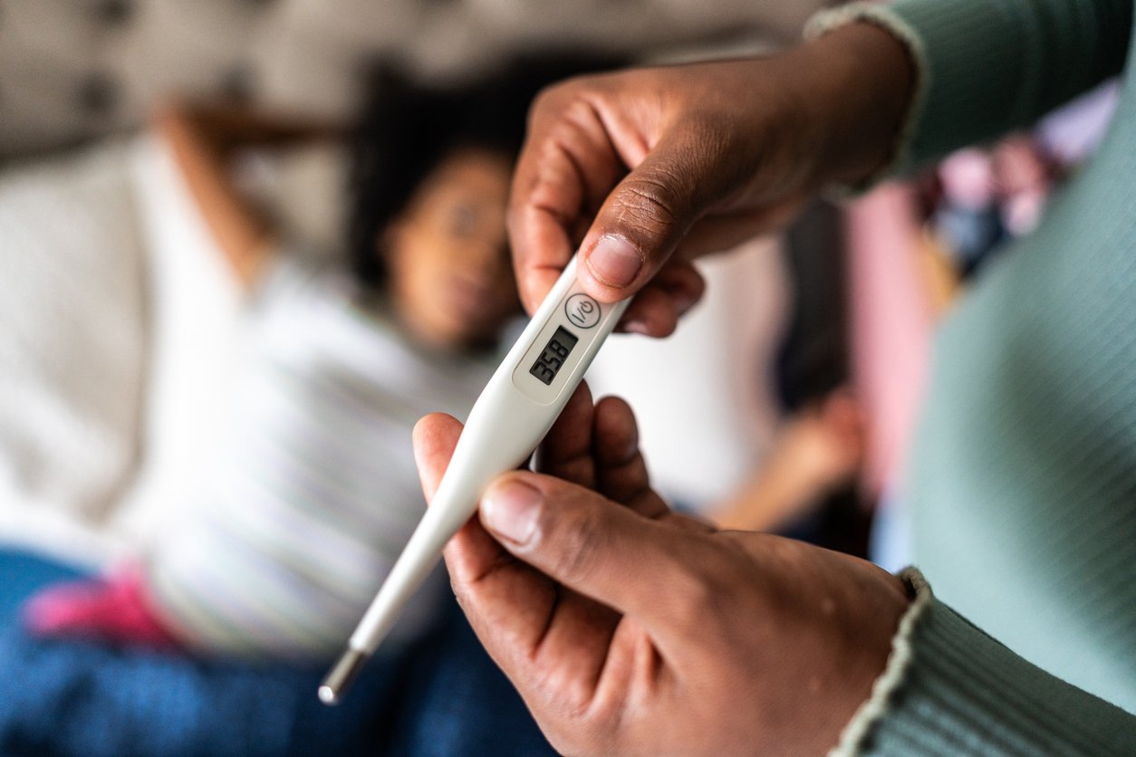 A close-up of hands holding a digital thermometer displaying a temperature of 38.8°C, with a person lying in bed in the blurred background.