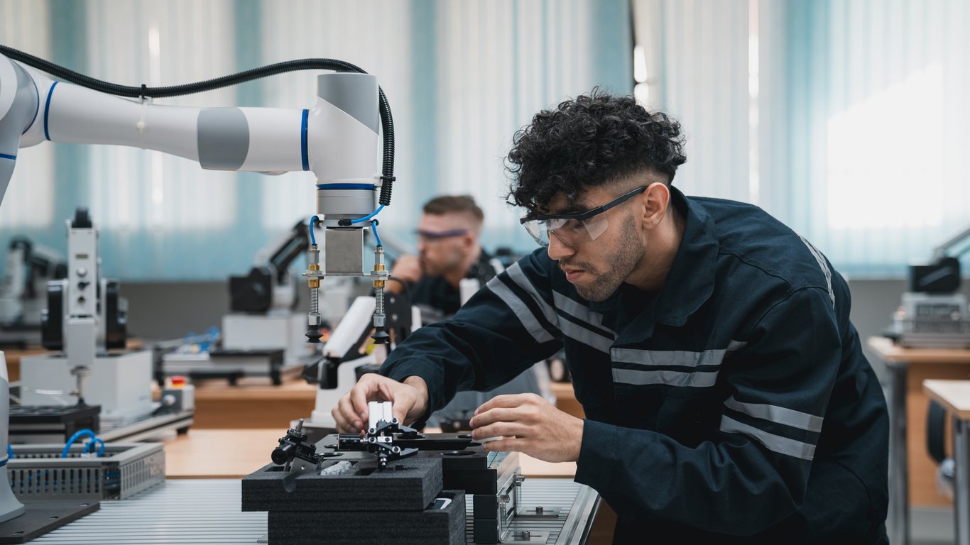 A man wearing safety glasses operates machinery next to a robotic arm in a modern, well-lit lab or factory setting. Another person works in the background.