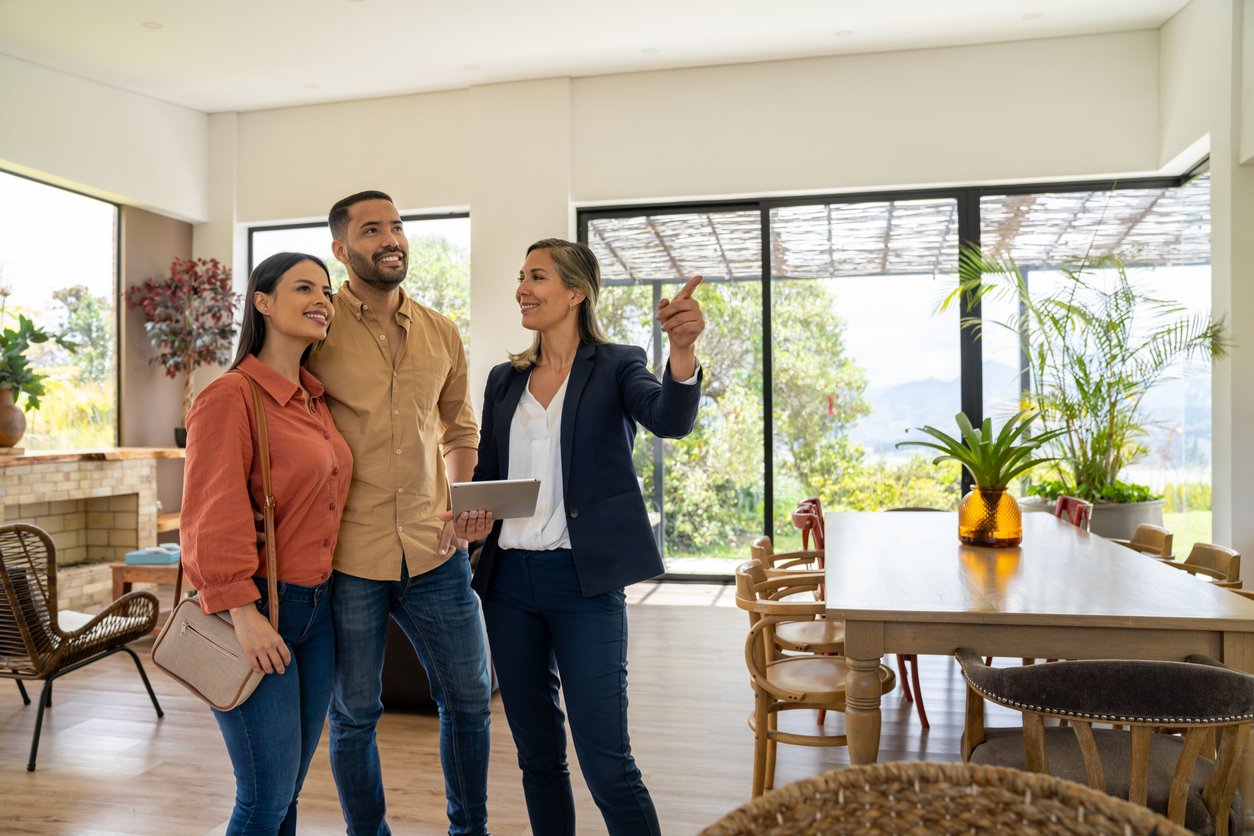 A real estate agent shows a smiling couple around a bright, modern home with large windows, a dining table, and indoor plants, while pointing out features in the open living space.