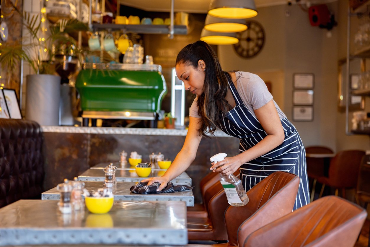 A woman wearing an apron cleans a table in a cozy café, holding a spray bottle and cloth. The café has a warm atmosphere with yellow lights, brown seating, and a green espresso machine in the background.