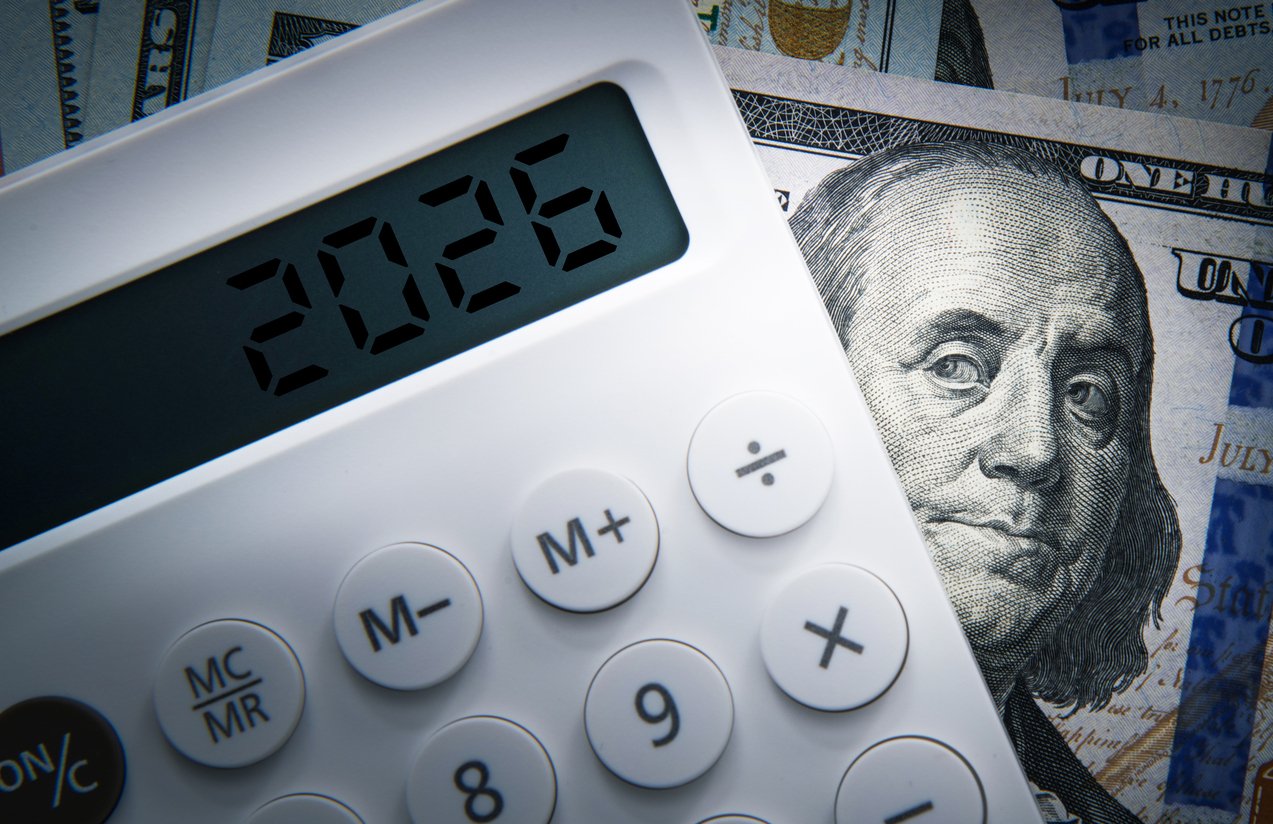 A white calculator displaying "2026" sits on top of US one hundred dollar bills, with Benjamin Franklin's face visible in the background.
