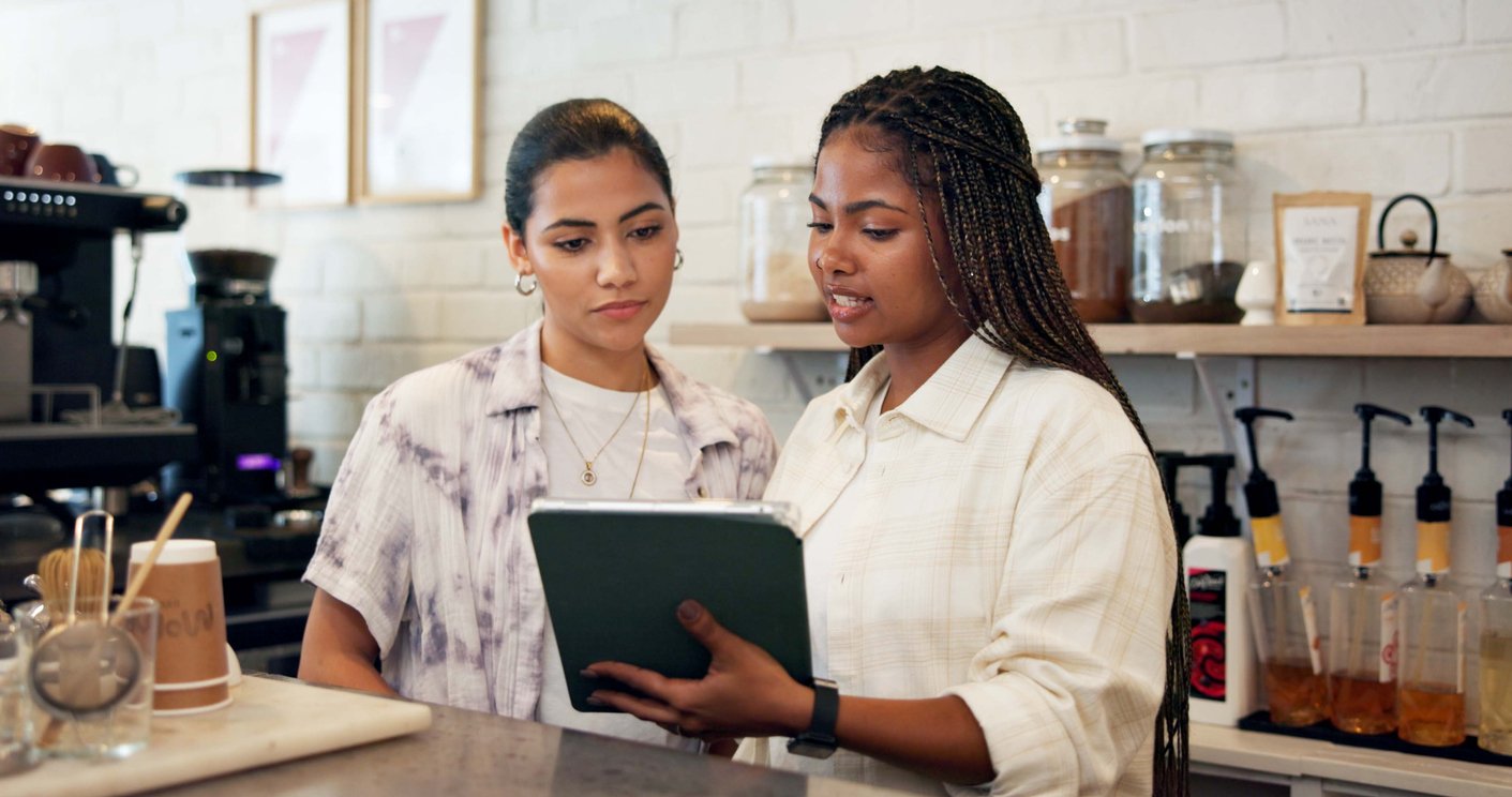 Two women stand behind a counter in a café, looking at a tablet together. One holds the tablet while the other watches. Shelves with jars and bottles are visible in the background.