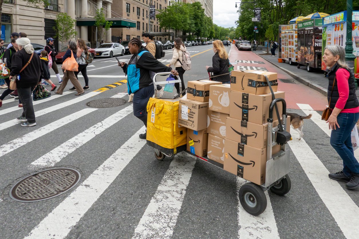 A delivery worker pushes a cart loaded with Amazon boxes across a busy crosswalk as people walk in different directions in an urban setting with buildings, trees, and food carts visible.