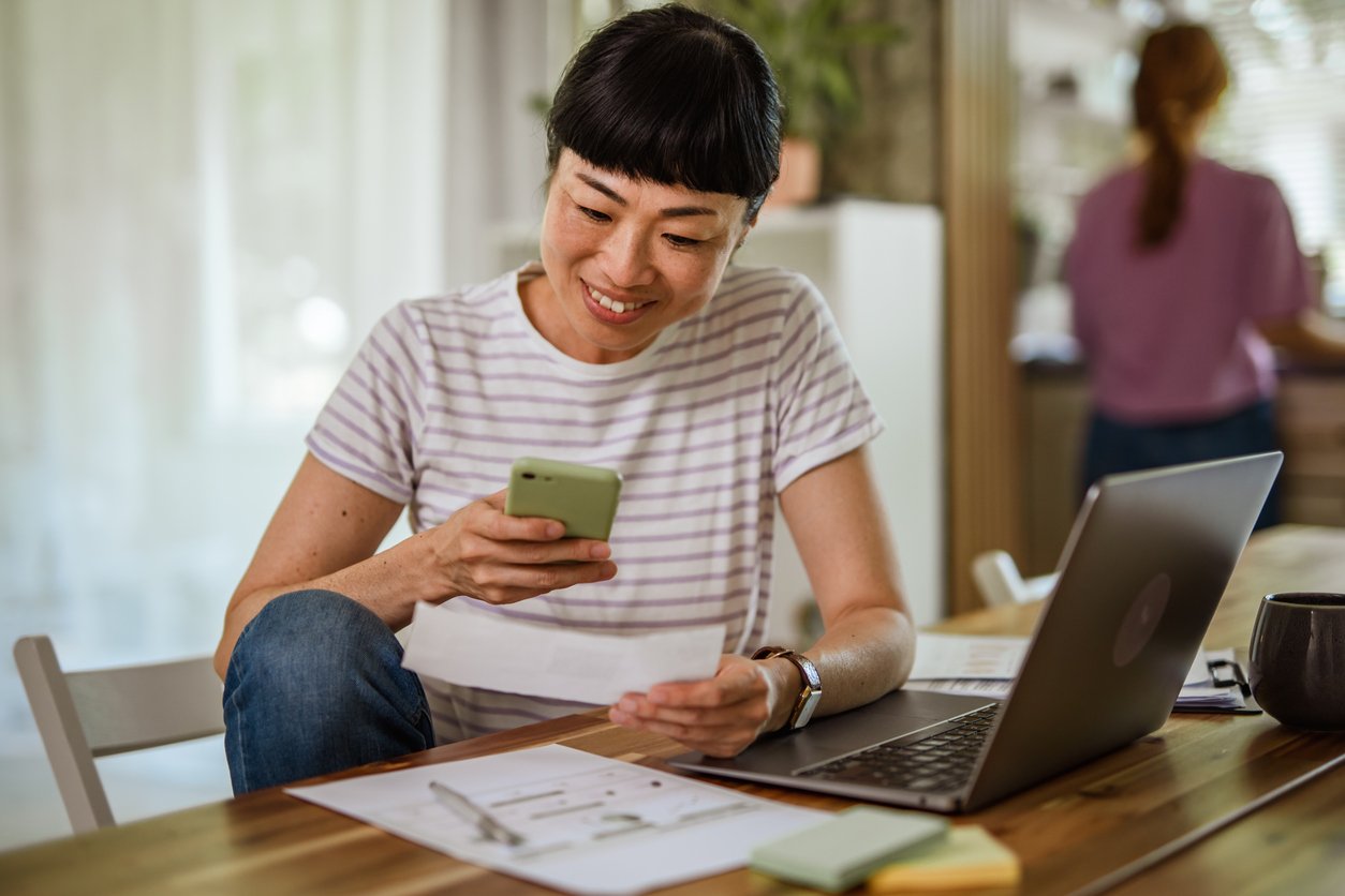A woman sits at a table, smiling while looking at her phone and holding a piece of paper. A laptop, documents, and a mug are on the table. Another person is blurred in the background.