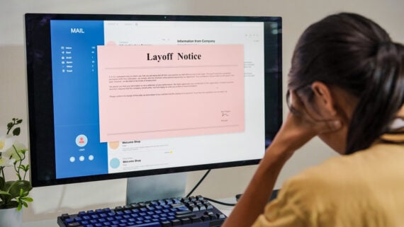 A person sits in front of a computer, looking upset, while reading a large "Layoff Notice" displayed on the screen, indicating job termination. An email inbox is visible in the background.