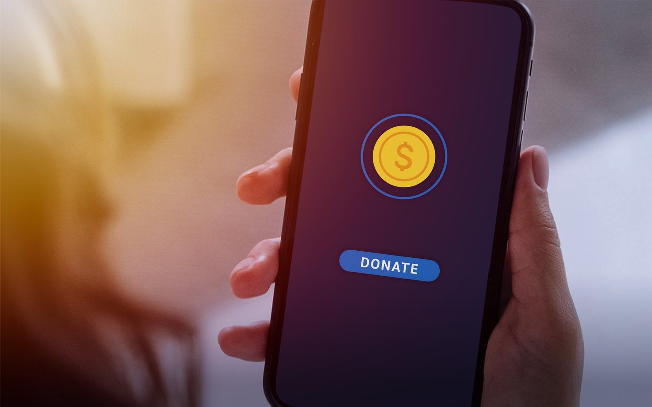 A person holds a smartphone displaying a screen with a gold coin symbol and a blue "DONATE" button, suggesting an online donation or charity app.