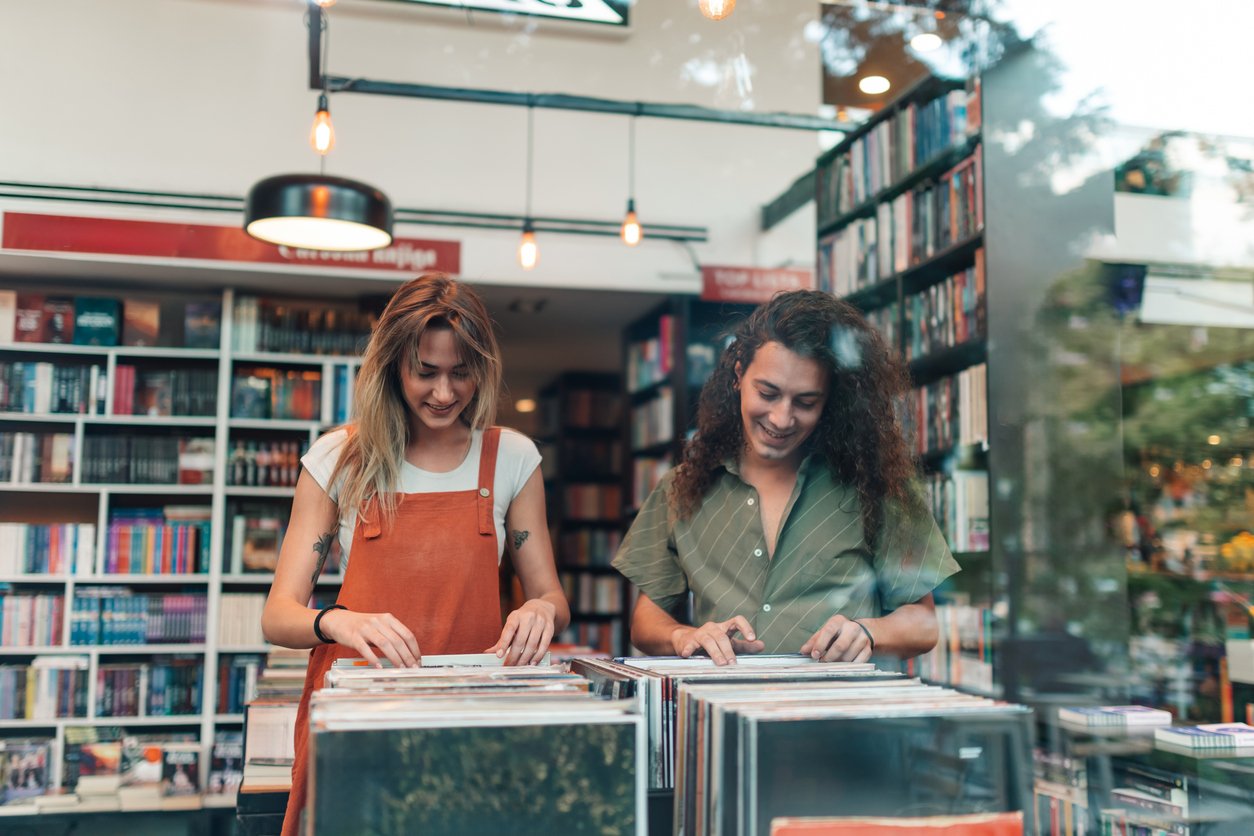 Two people browsing through records in a store, smiling and looking down at the vinyls. Bookshelves filled with books are visible in the background, and the scene is viewed through a glass window.