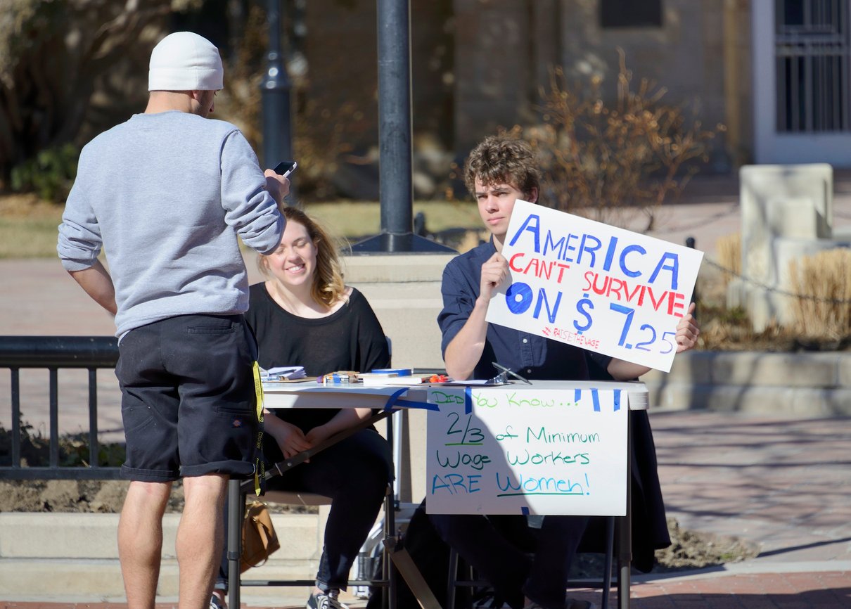 Two people sit at a table outside with signs about minimum wage. One sign reads “America can’t survive on $7.25.” A man in a beanie and shorts stands in front of the table, talking to them.