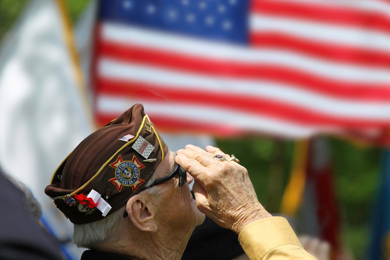An elderly veteran wearing a decorated cap salutes in front of a large, unfurled American flag during a ceremony.