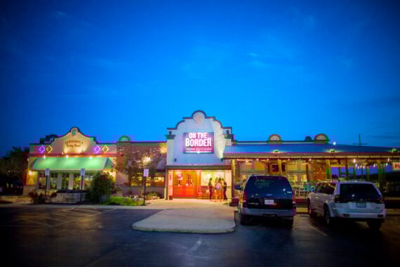 The exterior of an "On The Border" Mexican restaurant at dusk, with its neon sign lit, two people standing at the entrance, and several cars parked in the parking lot. The sky is a vibrant blue.