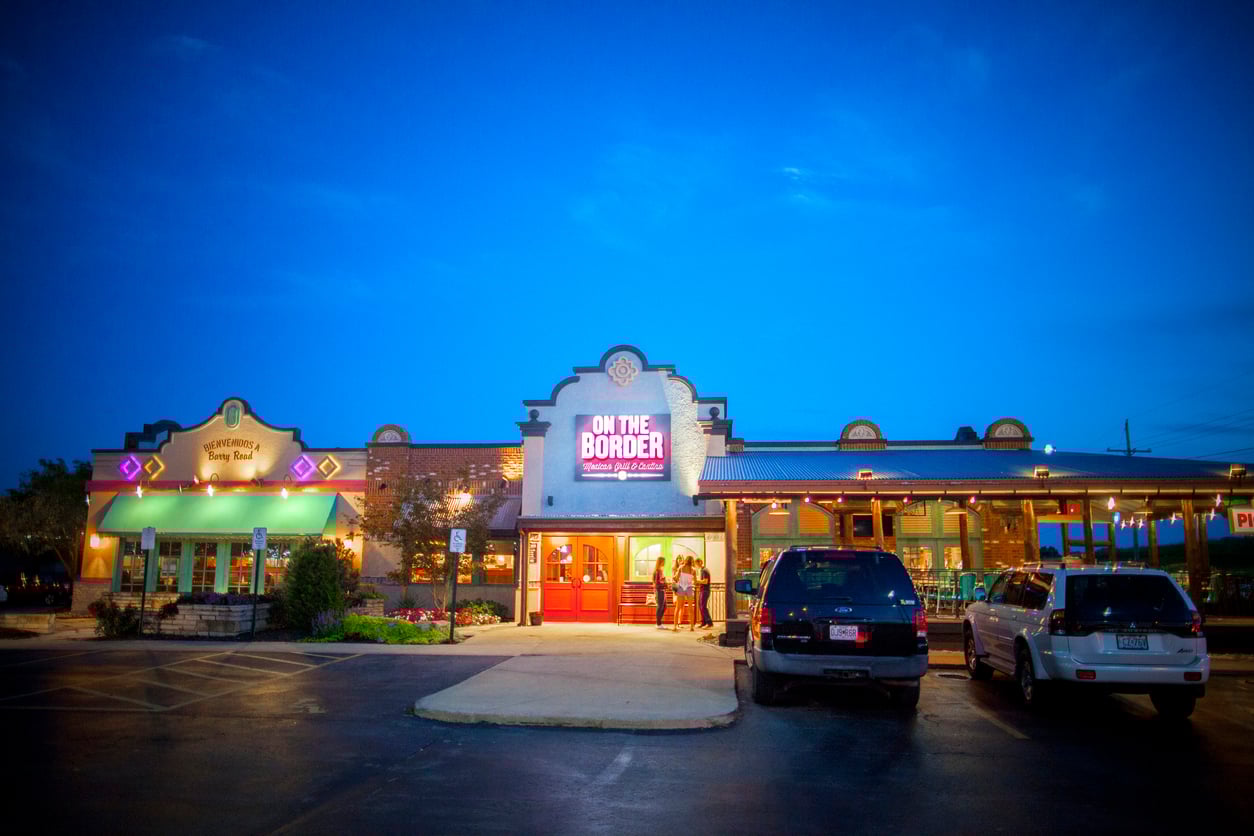 The exterior of an "On The Border" Mexican restaurant at dusk, with its neon sign lit, two people standing at the entrance, and several cars parked in the parking lot. The sky is a vibrant blue.