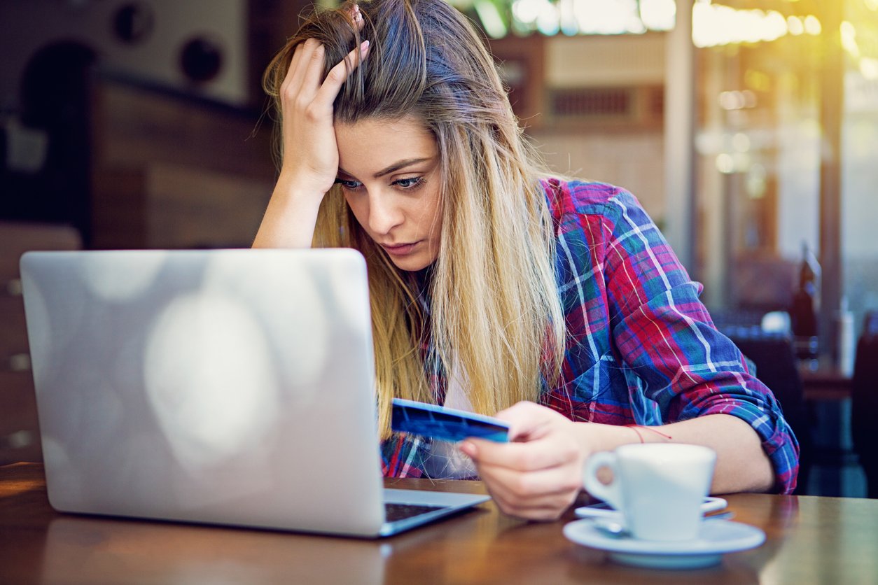 A woman with long blonde hair looks stressed while holding a credit card in one hand and touching her forehead with the other, staring at a laptop screen in a café with a coffee cup in front of her.