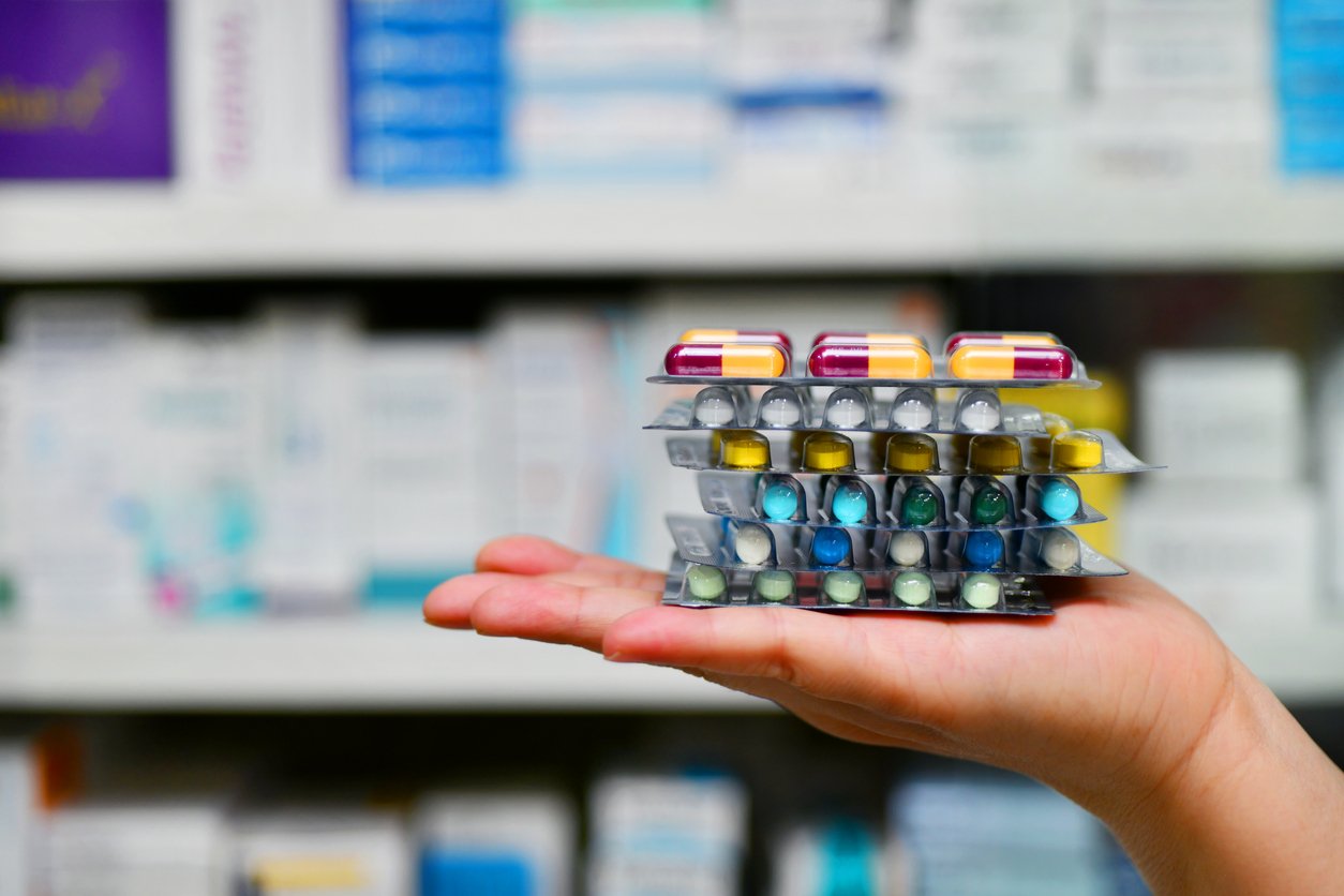 A hand holds several stacked blister packs of assorted pills in front of blurred pharmacy shelves stocked with various boxes of medication.